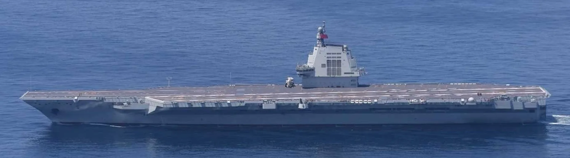 Chinese aircraft carrier Fujian sailing on a blue sea seen side-on, with its island superstructure near the stern