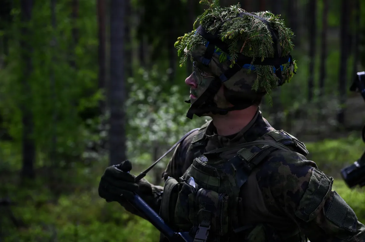 A Finnish soldier in face paint and a foliage-covered helmet holds a rifle in a pine forest