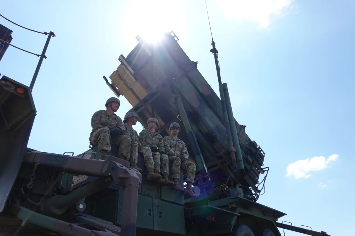 Four US soldiers sit on a Patriot missile launcher, its canisters raised against a bright blue sky