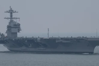 Grey US Navy aircraft carrier John F. Kennedy sailing in hazy weather with crew lining the flight deck