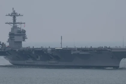 Grey US Navy aircraft carrier John F. Kennedy sailing in hazy weather with crew lining the flight deck