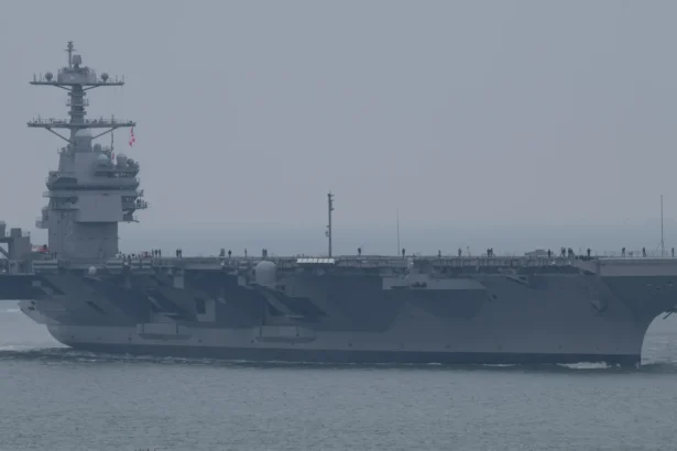 Grey US Navy aircraft carrier John F. Kennedy sailing in hazy weather with crew lining the flight deck